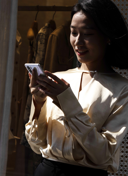 A person browsing on a smartphone in front of a clothing rack, symbolising consumer spending behaviour and modern shopping habits. Ideal for illustrating consumer spending trends, consumer spending reports, and consumer spending statistics.